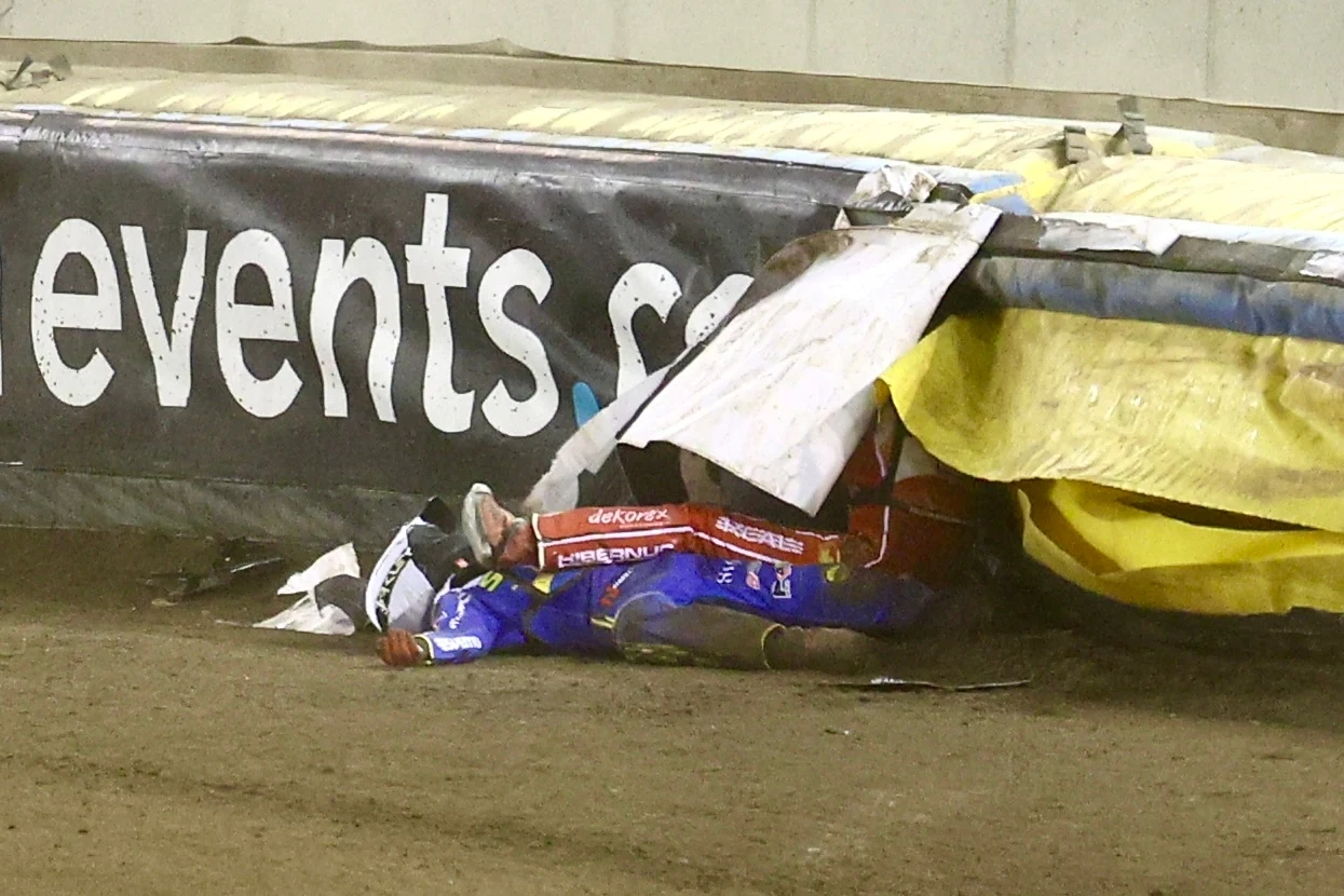 Zawodnik żużlowy w kombinezonie i kasku leży na torze częściowo pod bandą ochronną po upadku podczas wyścigu, w tle widoczna część reklamy na bandzie.