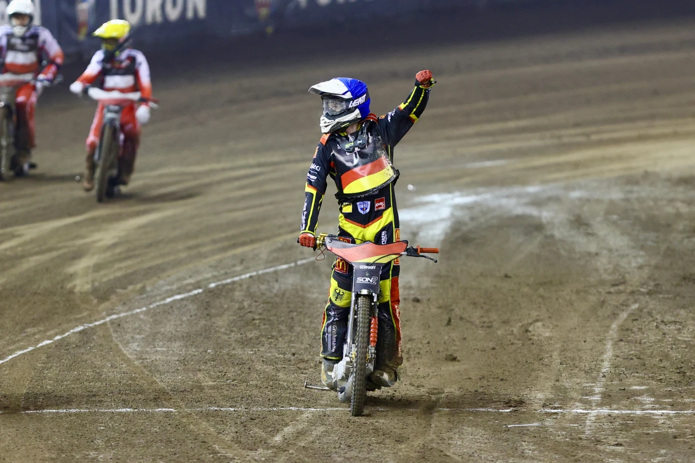 Zawodnik na motocyklu żużlowym podnosi rękę w geście triumfu, jadąc po torze, w tle inni zawodnicy oraz wyraźnie widoczna nawierzchnia toru żużlowego.