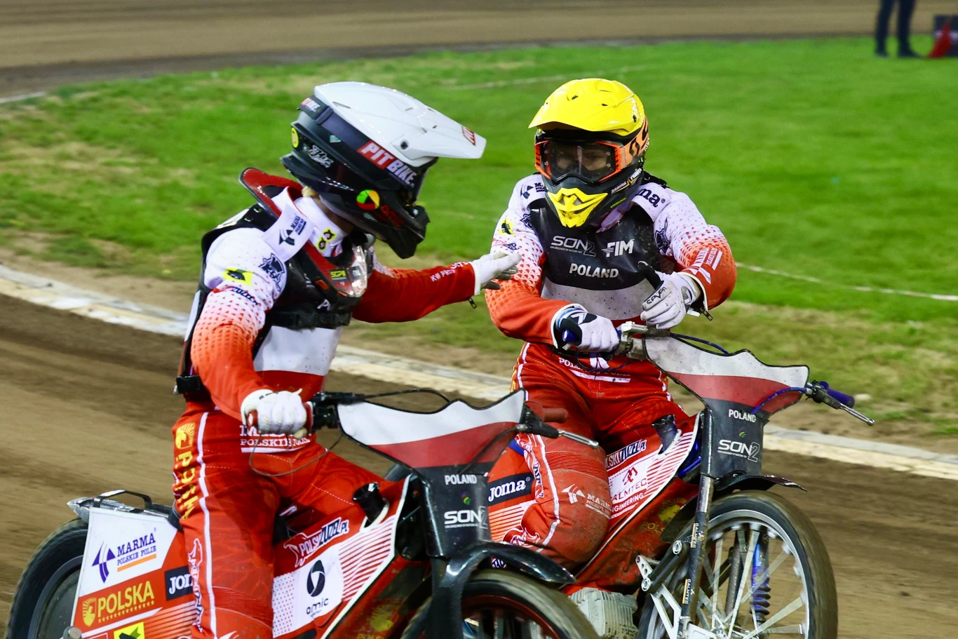 Dwaj żużlowcy w czerwonych kombinezonach i kaskach jadą obok siebie na torze żużlowym, wykonują gest wzajemnego wsparcia, wokół nich znajduje się zielona murawa i fragment stadionu.