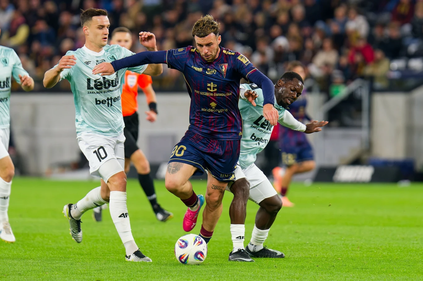 Piłkarz w granatowo-bordowym stroju walczy o piłkę z dwoma zawodnikami w jasnych strojach podczas meczu na stadionie, w tle widoczna publiczność.