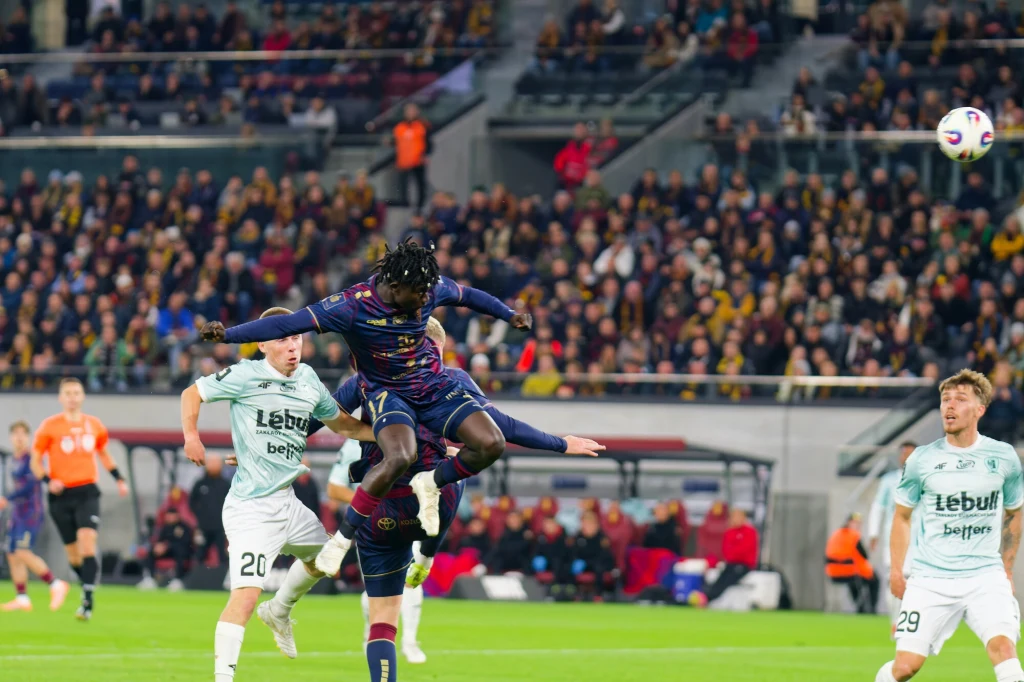 Pogoń Szczecin - Piast Gliwice Zawodnik w kolorowej koszulce wyskakuje w powietrze podczas akcji piłkarskiej otoczony przez rywali w jasnych strojach, w tle wypełnione trybuny i widoczna piłka.