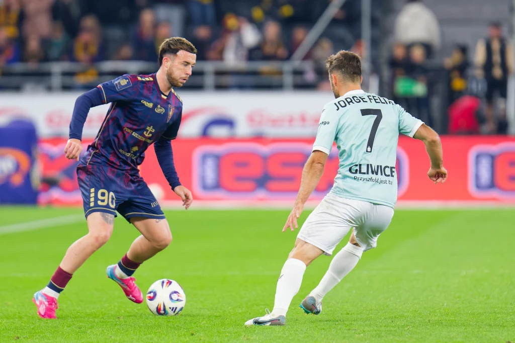 Sam Greenwood i Jorge Felix w meczu Pogoń Szczecin - Piast Gliwice Dwóch piłkarzy walczy o piłkę podczas meczu na profesjonalnym stadionie, jeden z nich ubrany w bordowo-granatowy strój, drugi w biały z numerem 7 i napisem 'Jorge Felix Gliwice', w tle widownia oraz reklamy.