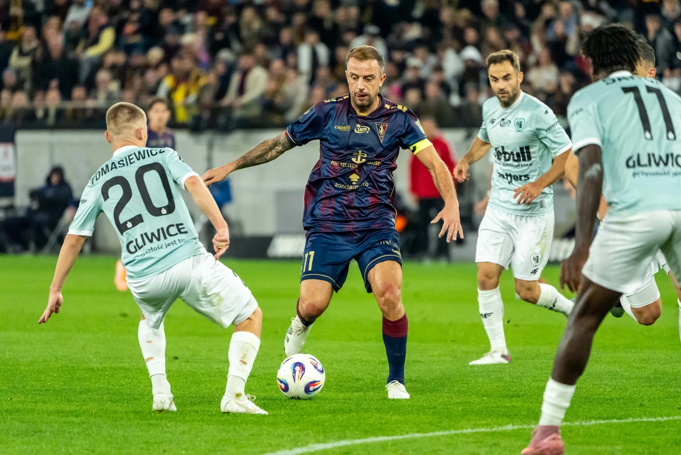 Kamil Grosicki w Pogoni Szczecin Piłkarze w trakcie meczu, zawodnik w granatowej koszulce prowadzi piłkę, otoczony przez zawodników w jasnych strojach, w tle widoczna publiczność na trybunach.