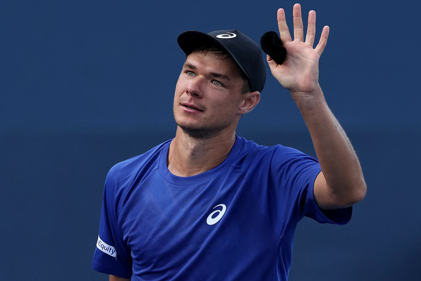 Mężczyzna w sportowym stroju i czapce z daszkiem unosi rękę z otwartą dłonią w geście pozdrowienia na tle niebieskiej ściany.