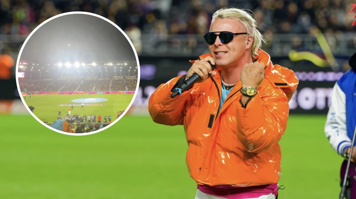 Wokalista w pomarańczowej kurtce i okularach przeciwsłonecznych występuje na stadionie, trzyma mikrofon i śpiewa do tłumu kibiców; w tle panorama stadionu z jasnymi światłami reflektorów i publicznością.