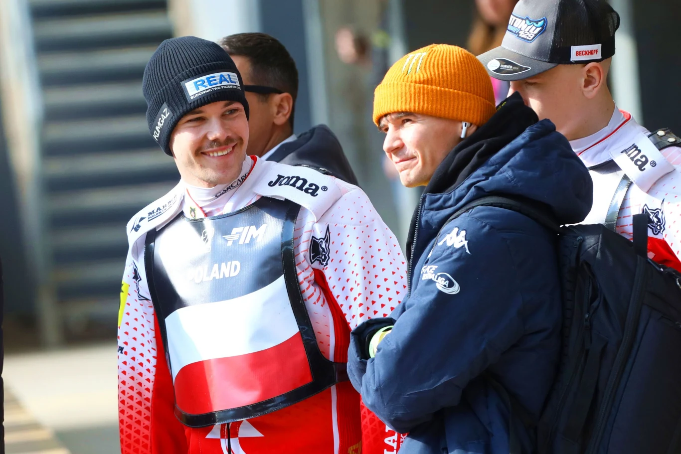 Młodzi mężczyźni ubrani w stroje sportowe z napisem POLAND i barwami narodowymi, stojący blisko siebie, rozmawiający i uśmiechnięci, jeden z nich ubrany w pomarańczową czapkę, luźna i przyjazna atmosfera.