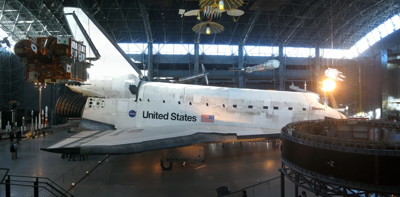 Wahadłowiec Discovery w Centrum Stevena F. Udvara-Hazy'ego Biały prom kosmiczny NASA z napisem United States oraz flagą Stanów Zjednoczonych ustawiony w dużej, nowoczesnej hali muzealnej, otoczony innymi elementami ekspozycji związanymi z technologią kosmiczną.
