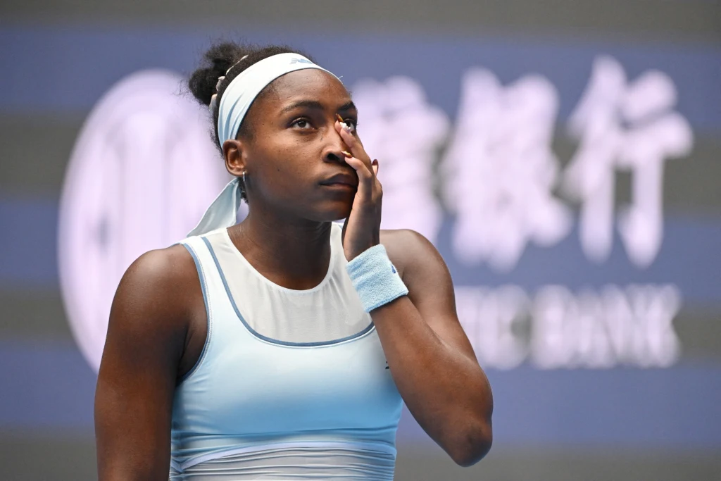 Tenisistka w błękitnym stroju sportowym stoi na korcie i dotyka twarzy, sprawiając wrażenie zamyślonej lub zmęczonej, w tle widoczne rozmyte logo banku.