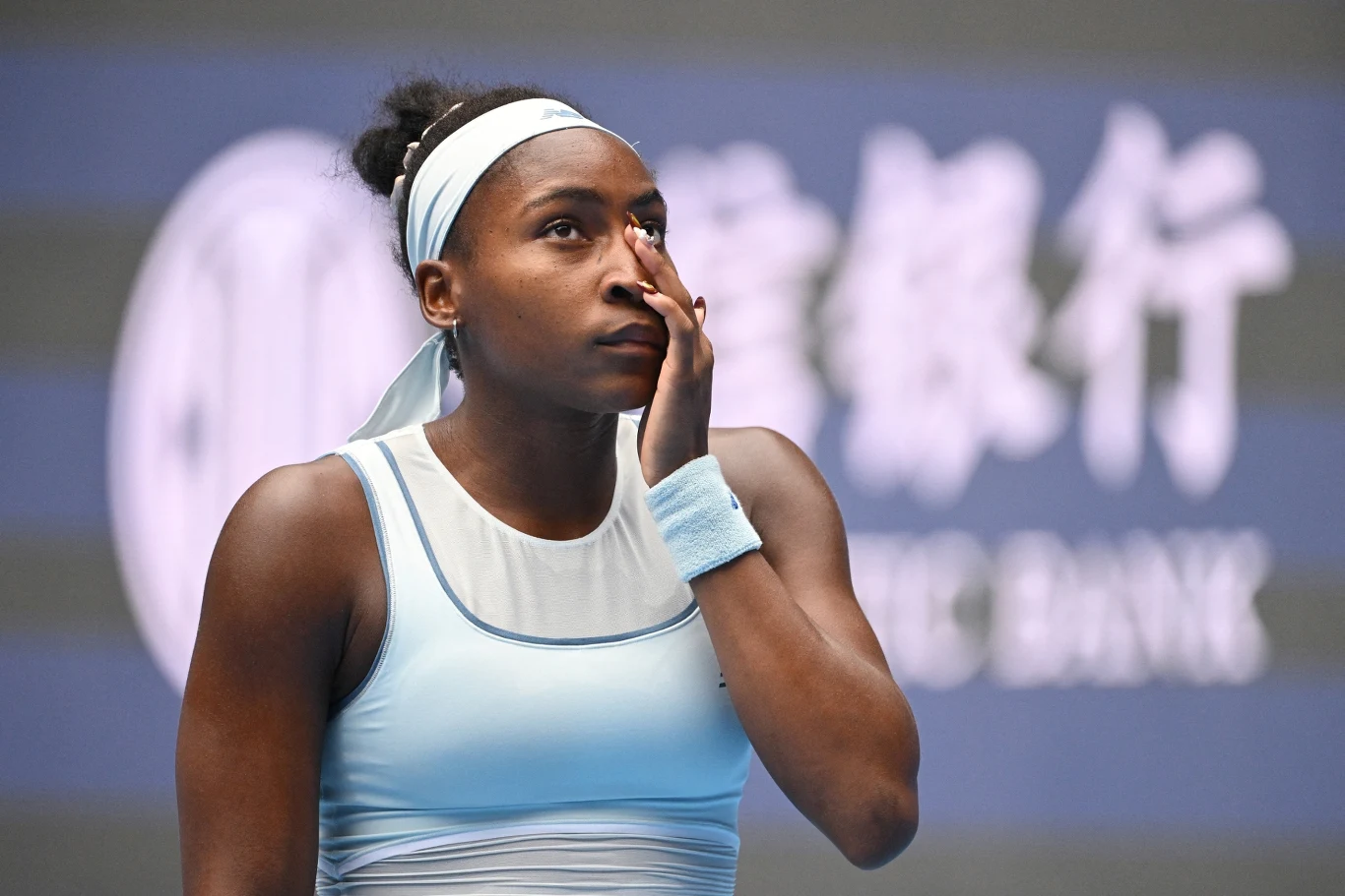 Tenisistka w błękitnym stroju sportowym stoi na korcie i dotyka twarzy, sprawiając wrażenie zamyślonej lub zmęczonej, w tle widoczne rozmyte logo banku.