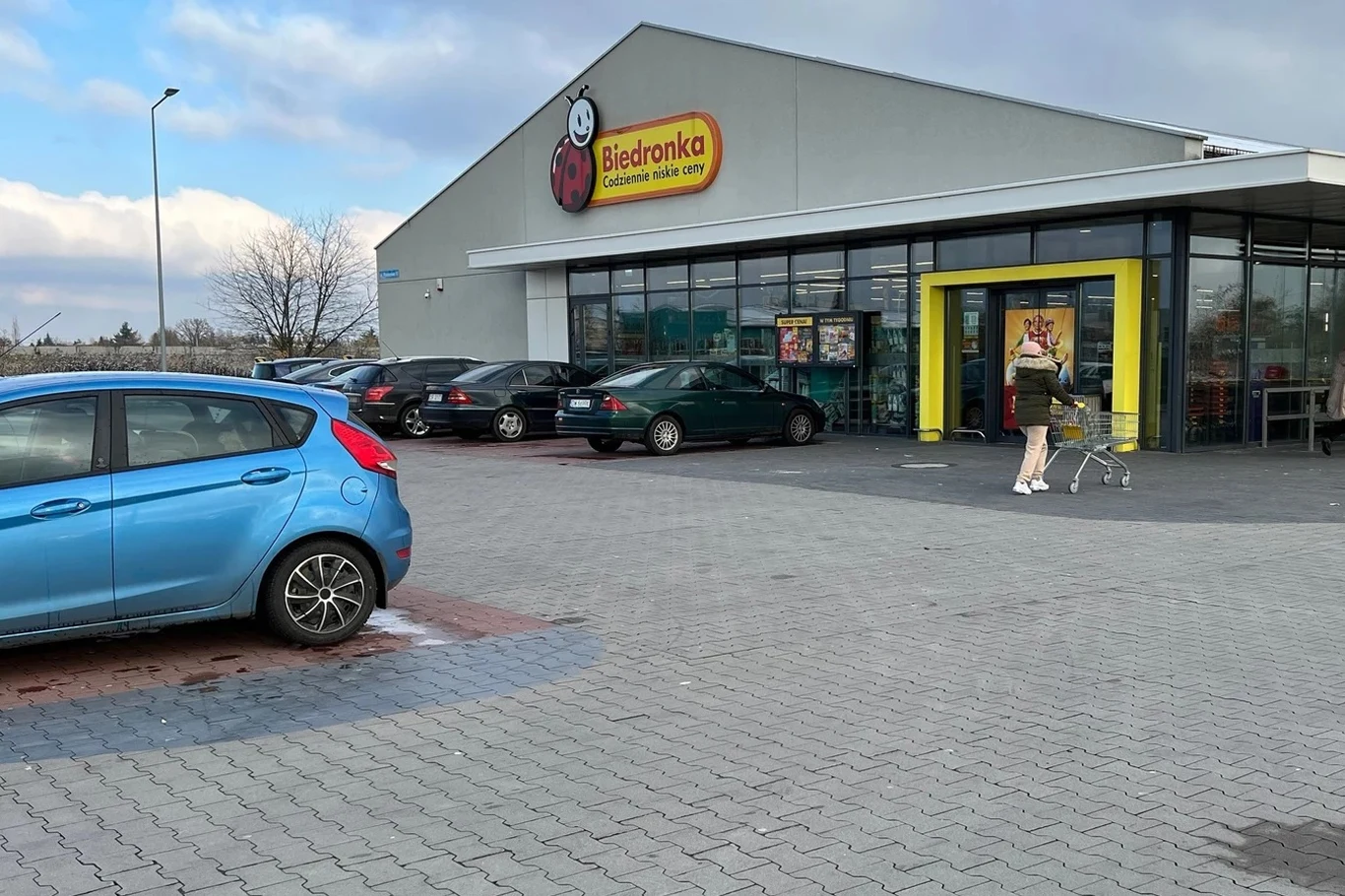 Biedronka testuje system automatycznego naliczania opłat na swoich parkingach Parking przed supermarketem Biedronka z zaparkowanymi samochodami i osobą pchającą wózek sklepowy w kierunku wejścia do budynku.
