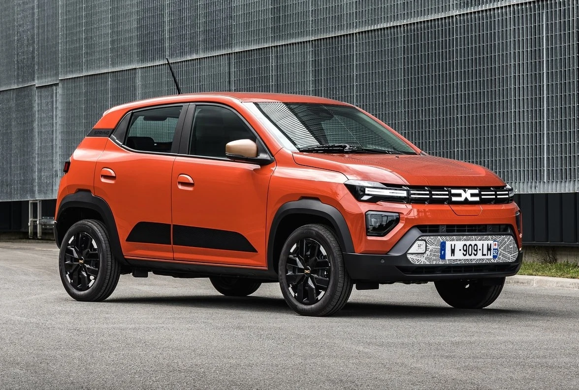 Dacia Spring to obecnie najtańszy samochód elektryczny w Polsce Nowoczesny, pomarańczowy samochód typu SUV z czarnymi elementami nadwozia i czarnymi felgami stoi na asfalcie przed metalowym ogrodzeniem.