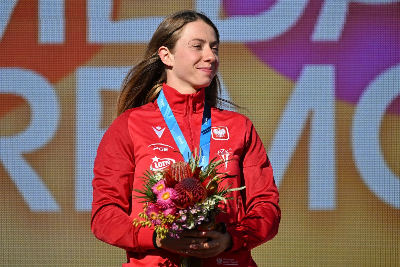 Klaudia Zwolińska Sportowiec w czerwonym dresie reprezentacji Polski stoi na tle kolorowej planszy z medalem na szyi i bukietem kwiatów w dłoniach