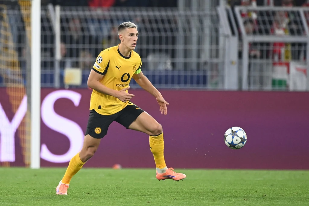 Nico Schlotterbeck Piłkarz w żółto-czarnym stroju klubu Borussia Dortmund biegnie z piłką po murawie stadionu, w tle widoczne trybuny i ogrodzenie.