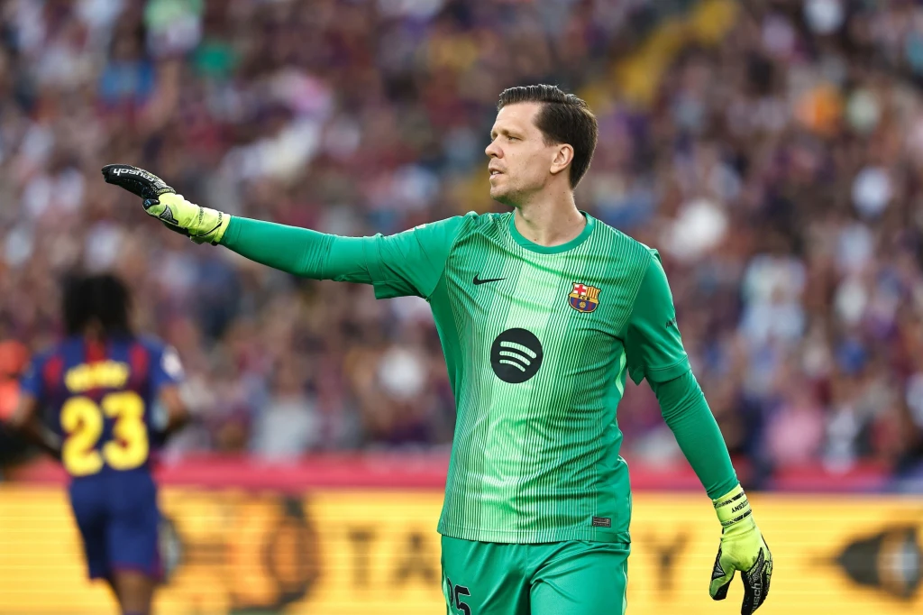 Bramkarz w zielonym stroju piłkarskim pokazuje kierunek z wyciągniętą ręką, na tle stadionu pełnego kibiców, drugi zawodnik w żółto-czerwonym stroju widoczny w rozmyciu.