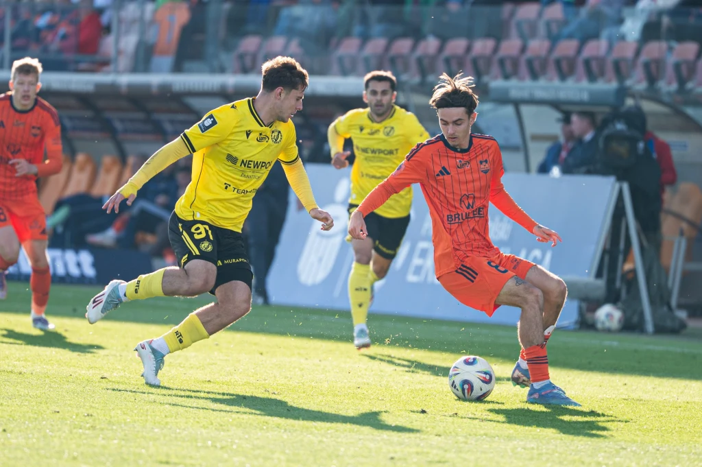 Mecz Termalica - Widzew Łódź Dwóch piłkarzy rywalizuje o piłkę podczas meczu piłki nożnej, jeden z nich ubrany w żółty strój, drugi w pomarańczowy, w tle widoczna trybuna z kibicami.