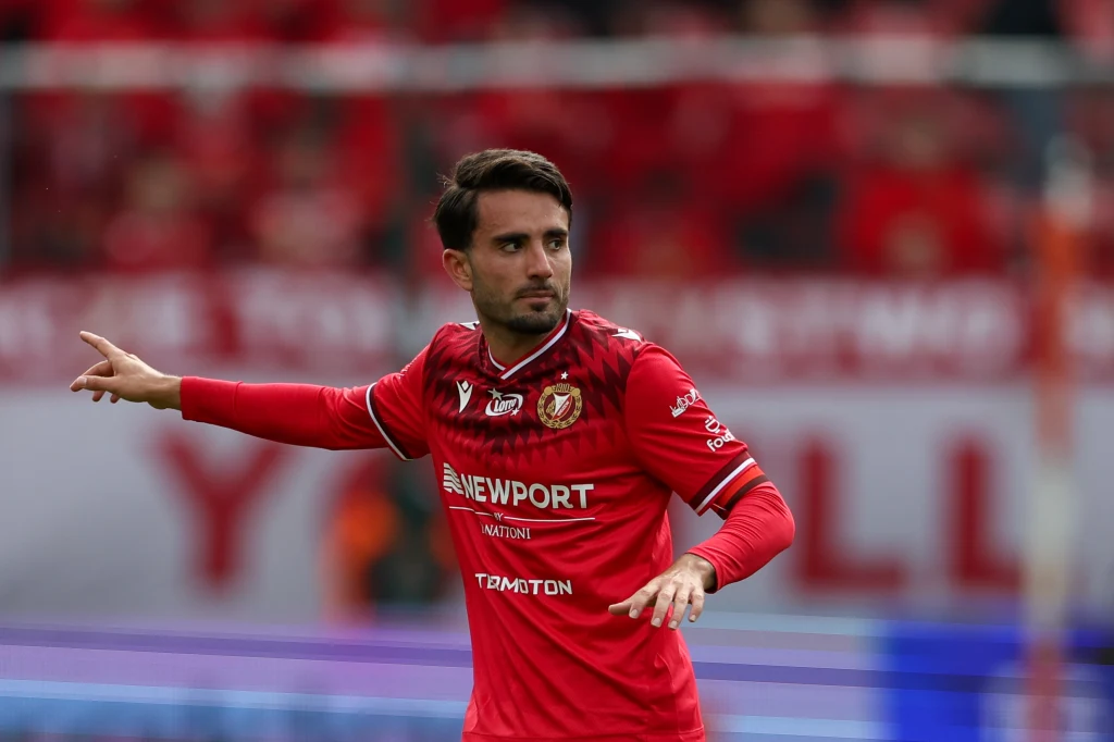 Fran Alvarez Piłkarz w czerwonym stroju z logo sponsorów na koszulce wykonuje gest ręką podczas meczu na stadionie, w tle niewyraźni kibice i trybuny pokryte czerwonymi barwami.