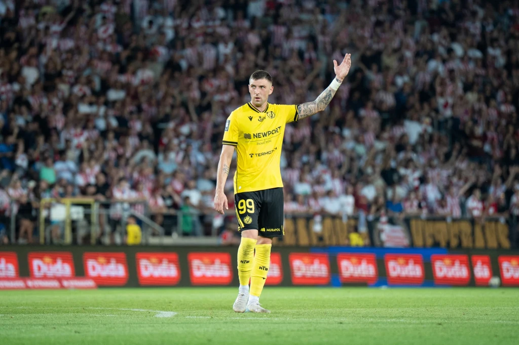 Sebastian Bergier Piłkarz w żółtej koszulce z numerem 99 na stadionie unoszący rękę, w tle trybuny pełne kibiców i reklamy na bandach.