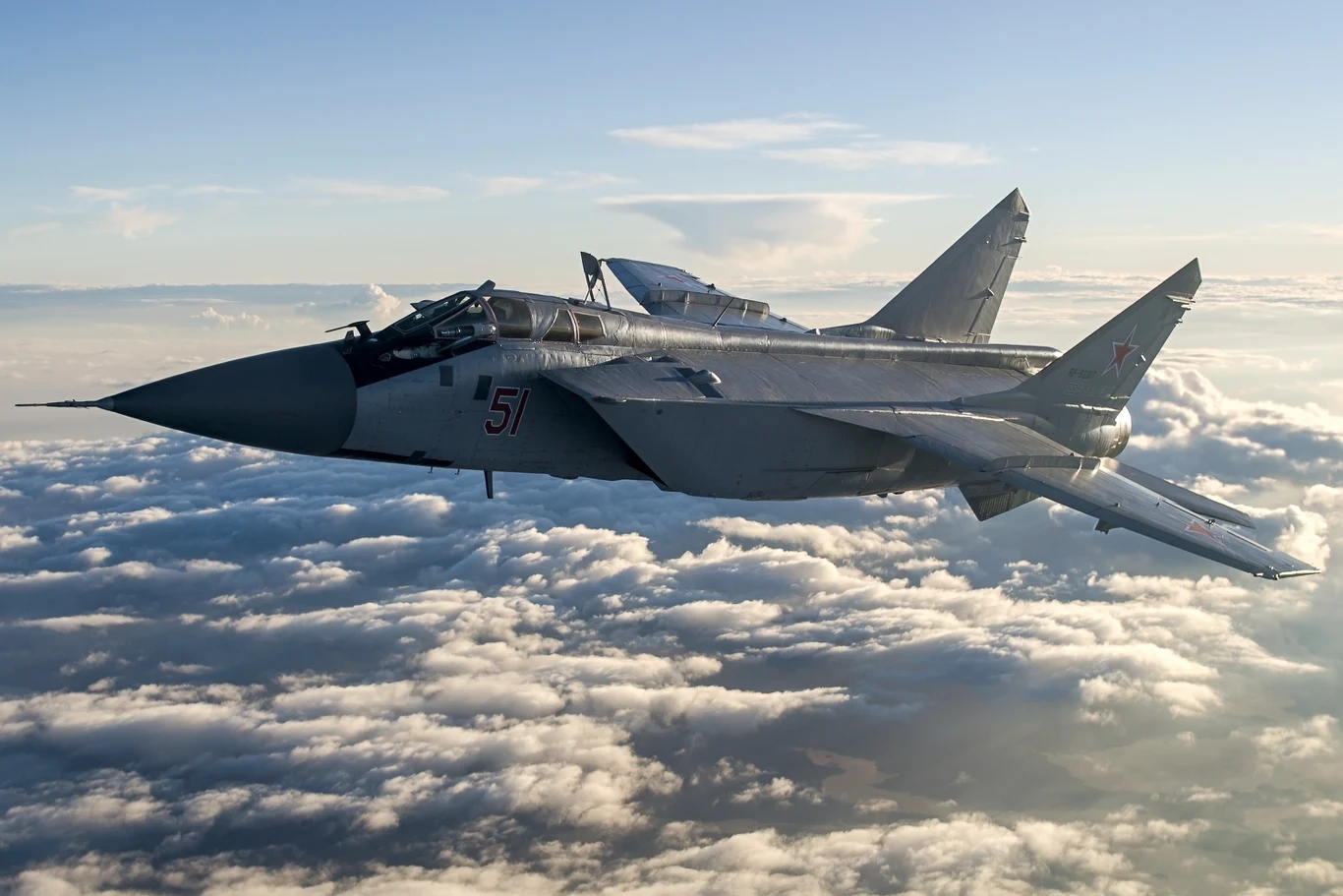 Myśliwiec wojskowy typu MiG-31 przelatuje nad pokrytymi chmurami, dobrze oświetlony przez słońce, widoczne są szczegóły konstrukcji maszyny, oznaczenia liczbowe i narodowe na stateczniku pionowym.