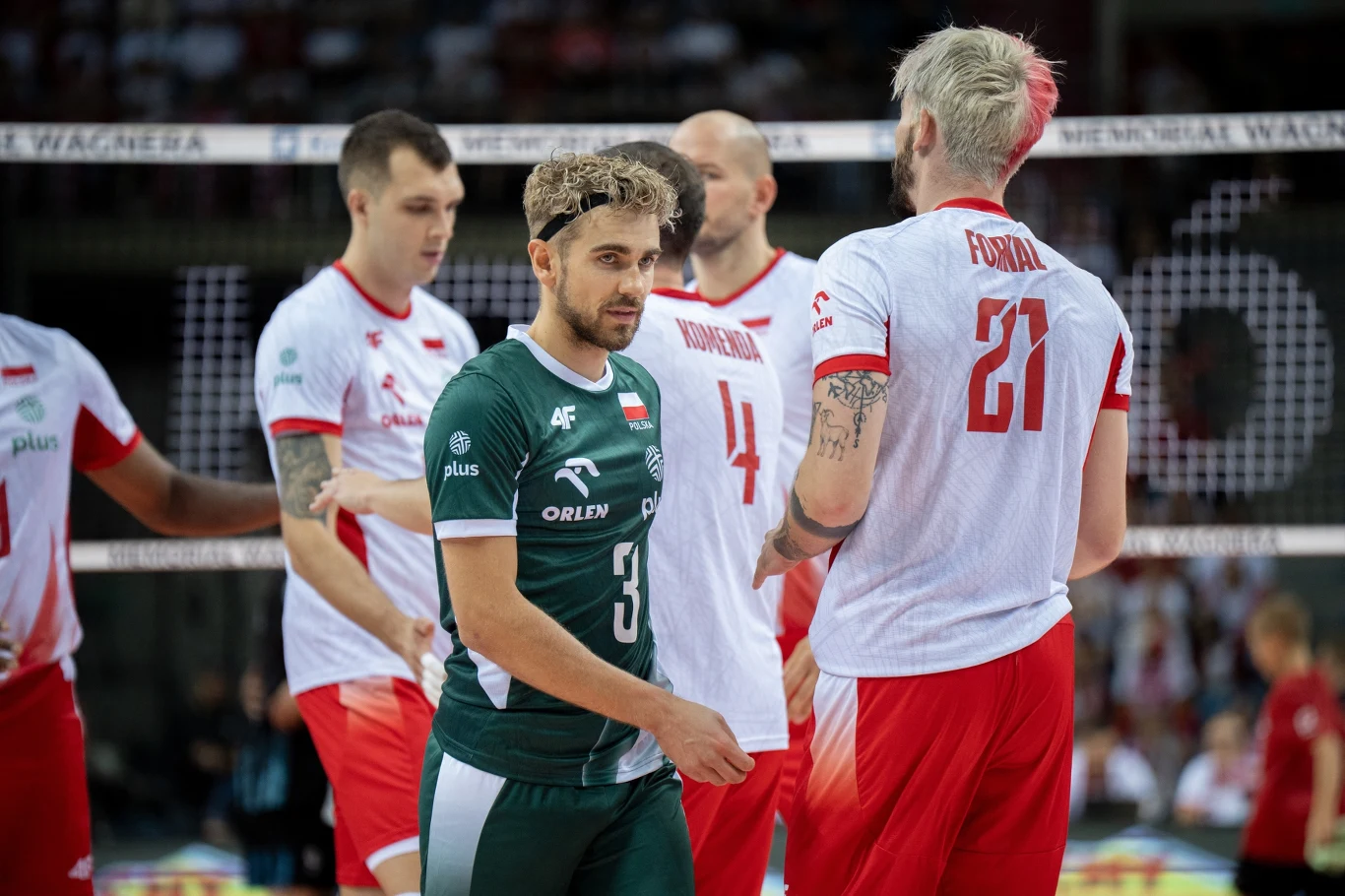 Grupa siatkarzy reprezentujących Polskę na hali podczas meczu, zawodnicy w białych i czerwonych strojach oraz jeden w zielonym stroju libero, intensywna atmosfera sportowej rywalizacji.