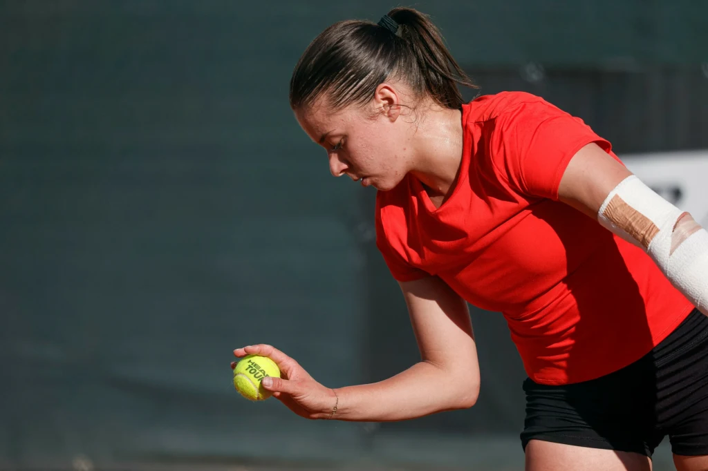 Maja Chwalińska Kobieta w czerwonej koszulce i czarnych spodenkach przygotowuje się do serwisu w tenisie, trzymając żółtą piłkę tenisową i pochylając się lekko do przodu; na jednym z jej łokci widoczny jest opatrunek.