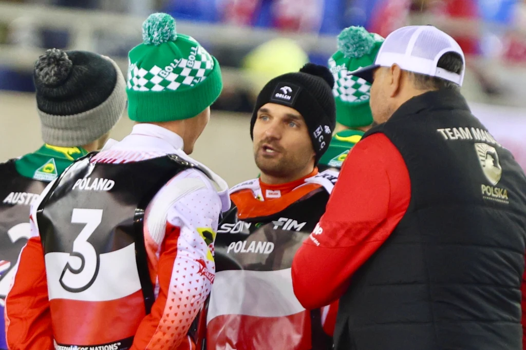 Dobrucki w rozmowie ze Zmarzlikiem i Pawlickim Grupa mężczyzn w sportowych strojach reprezentacji Polski i Australii rozmawia na stadionie, wszyscy ubrani w zimowe czapki i kurtki, widoczne napisy 'Poland', 'Australia' oraz logo drużyn narodowych.