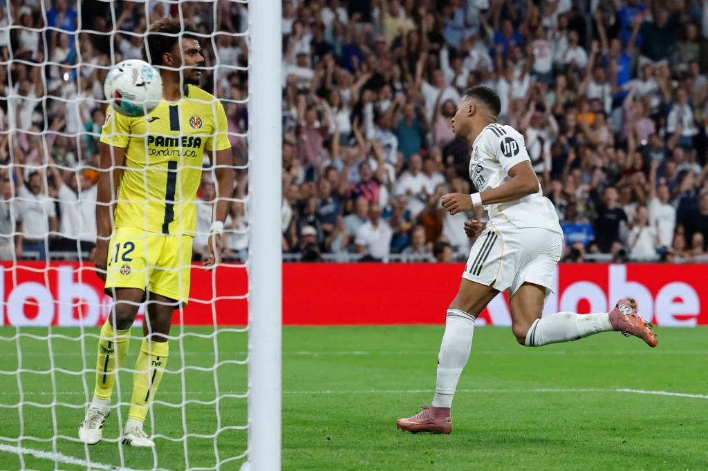 Real Madryt - Villarreal (4.10.2025) Piłkarz w białym stroju świętuje zdobycie gola, podczas gdy bramkarz w żółtym stroju trzyma piłkę, w tle widać rozemocjonowaną publiczność na stadionie.