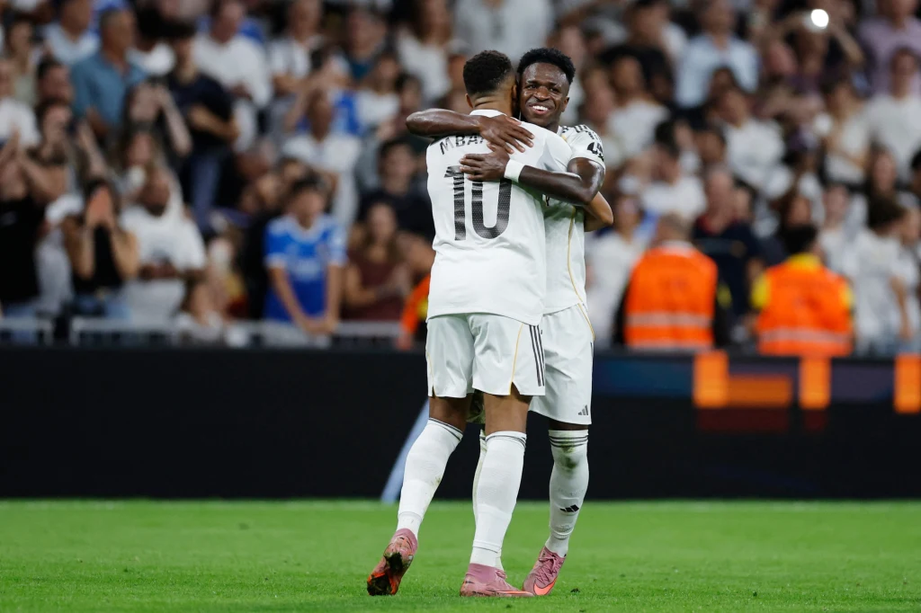 Kylian Mbappe i Vinicius Junior bohaterami meczu Realu Madryt z Villarrealem Dwóch piłkarzy w białych strojach obejmuje się na murawie stadionu, wokół nich tłum kibiców na trybunach oraz osoby w kamizelkach porządkowych w tle.