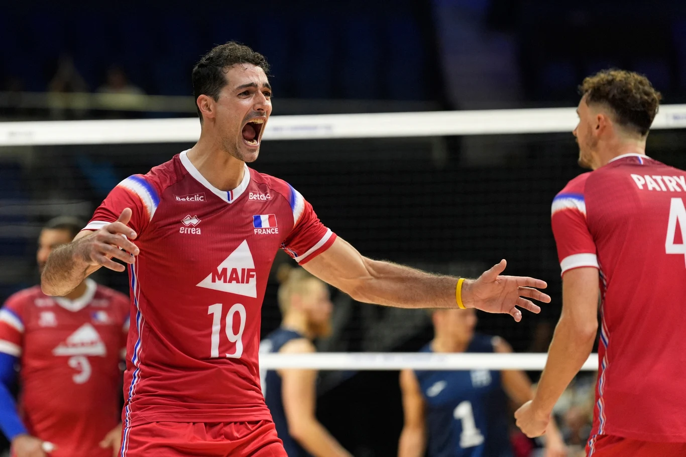 Grupa siatkarzy w czerwonych strojach świętująca punkt podczas meczu, zawodnik z numerem 19 ma szeroko rozłożone ramiona i wykrzykuje coś z radości, siatka w tle oddziela drużyny