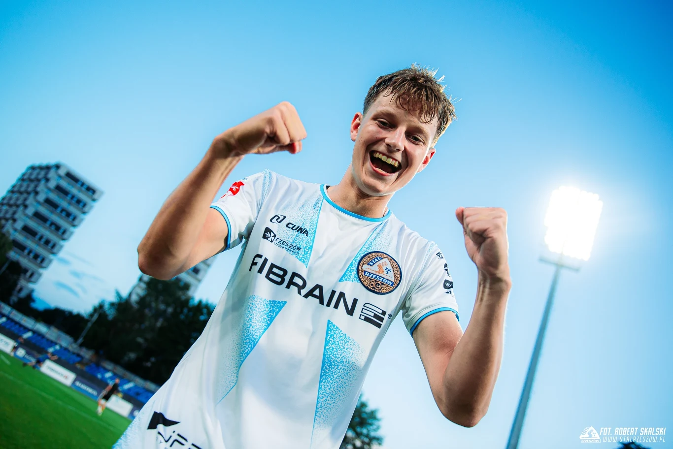 Młody piłkarz w białym stroju sportowym drużyny piłkarskiej świętuje sukces na stadionie, trzymając zaciśnięte pięści z szerokim uśmiechem na twarzy, w tle widać stadionowe światła i fragment trybun.