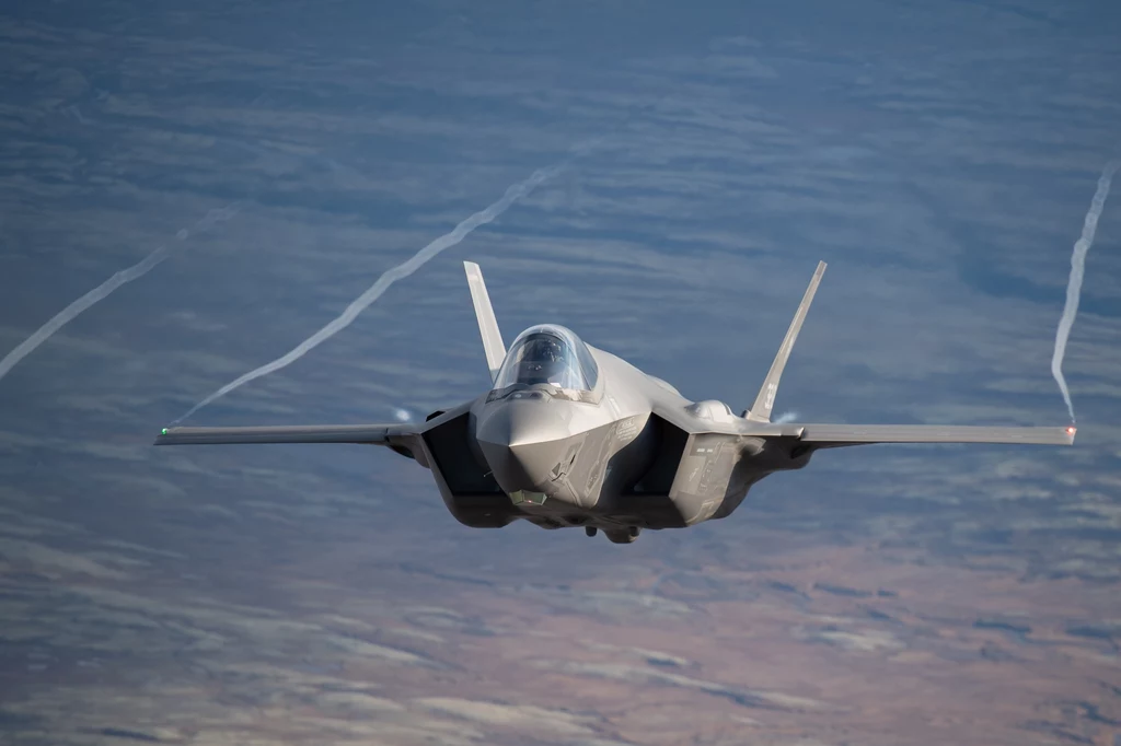 Nowoczesny myśliwiec wojskowy F-35 lecący na dużej wysokości nad rozległym terenem, smugami powietrznymi ciągnącymi się za skrzydłami.