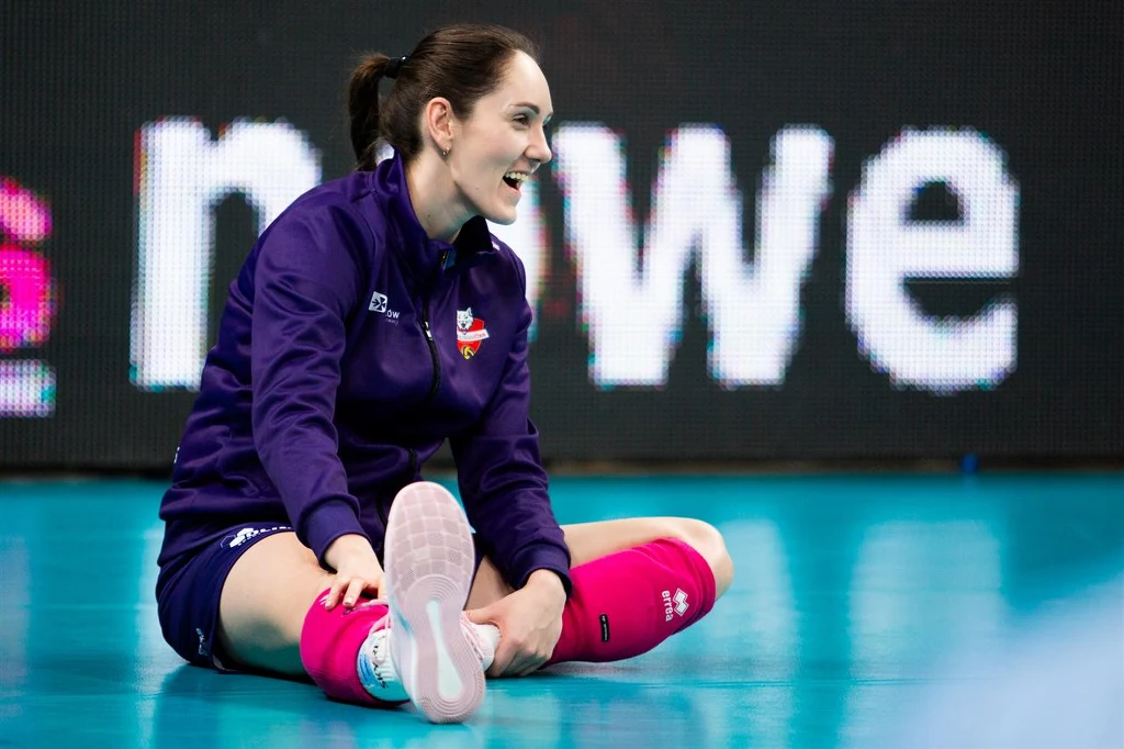 Katarzyna Wenerska, rozgrywająca DevelopResu Rzeszów i reprezentacji Polski Siatkarka ubrana w fioletową bluzę klubową i różowe getry siedzi na parkiecie sportowej hali z uśmiechem na twarzy, jedna noga wyciągnięta do przodu, w tle widoczne rozmazane elementy dekoracyjne boiska.