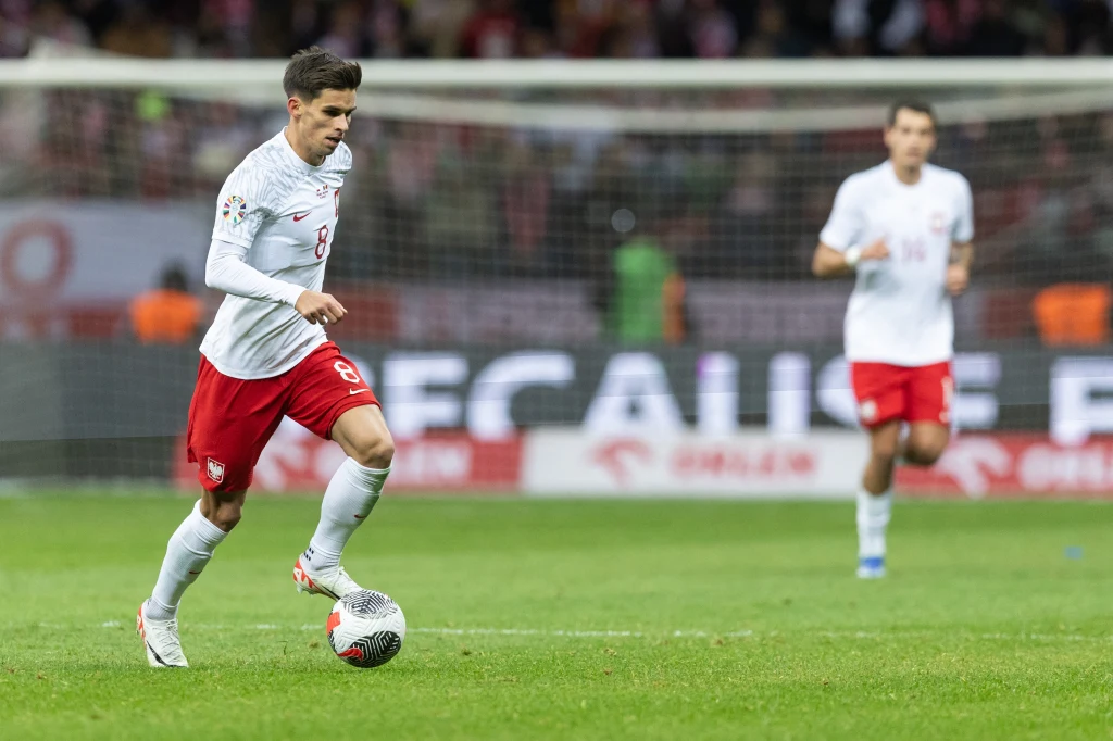 Piłkarz w białej koszulce i czerwonych spodenkach prowadzi piłkę po murawie stadionu podczas meczu, w tle widać drugiego zawodnika tej samej drużyny oraz bramkę.