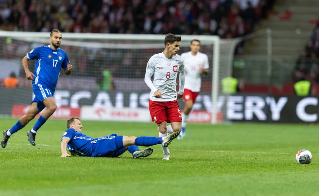 Zawodnik w biało-czerwonym stroju wykonuje zagranie piłką podczas meczu piłkarskiego, w tle dwóch zawodników drużyny przeciwnej w niebieskich strojach, jeden z nich leży na murawie po nieudanej próbie zatrzymania akcji.