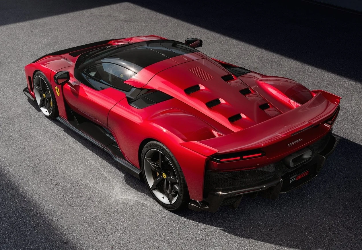 Luksusowy sportowy samochód marki Ferrari w intensywnym czerwonym kolorze, widok z góry na tylną część pojazdu z widocznymi aerodynamicznymi przetłoczeniami i charakterystycznym logo. Auto stoi na jasnej nawierzchni w dobrze oświetlonym miejscu.