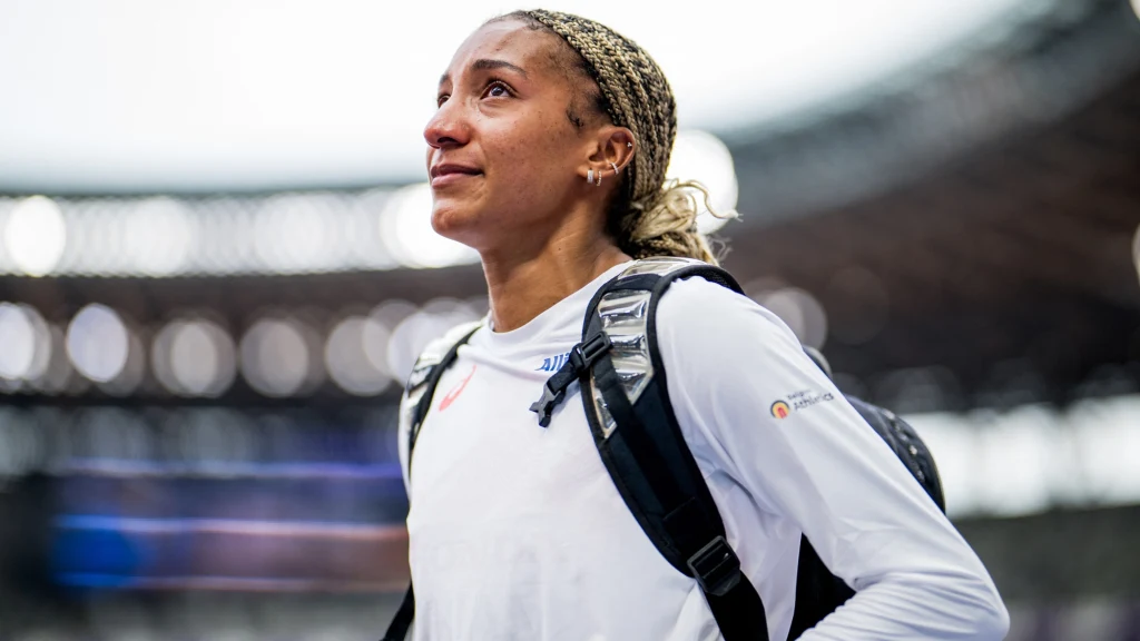 Nafissatou Thiam Kobieta ubrana w białą bluzę sportową z plecakiem na ramionach, patrząca w górę z delikatnym uśmiechem, w tle rozmazane światła stadionu.