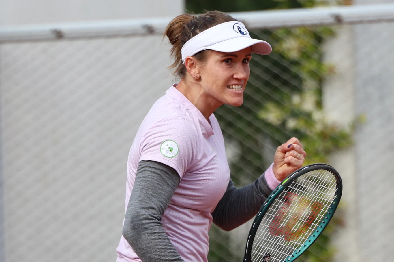 Kobieta ubrana w sportową odzież i czapkę z daszkiem ściska rękę w geście triumfu, trzymając rakietę tenisową, z wyraźną determinacją na twarzy na tle siatki tenisowej.