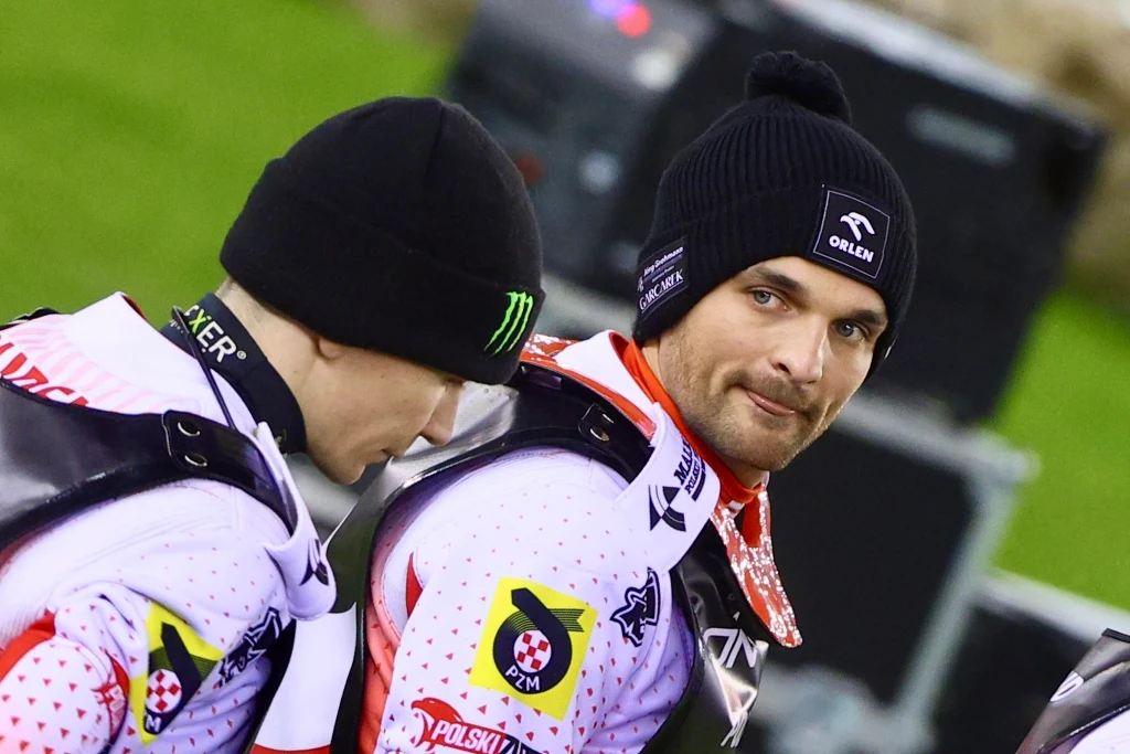 Bartosz Zmarzlik i Patryk Dudek Dwóch żużlowców w białych strojach sportowych, ubrani w czarne czapki, stoją obok siebie na stadionie, na ich kombinezonach widoczne są logotypy sponsorów, jeden z nich patrzy w stronę obiektywu.