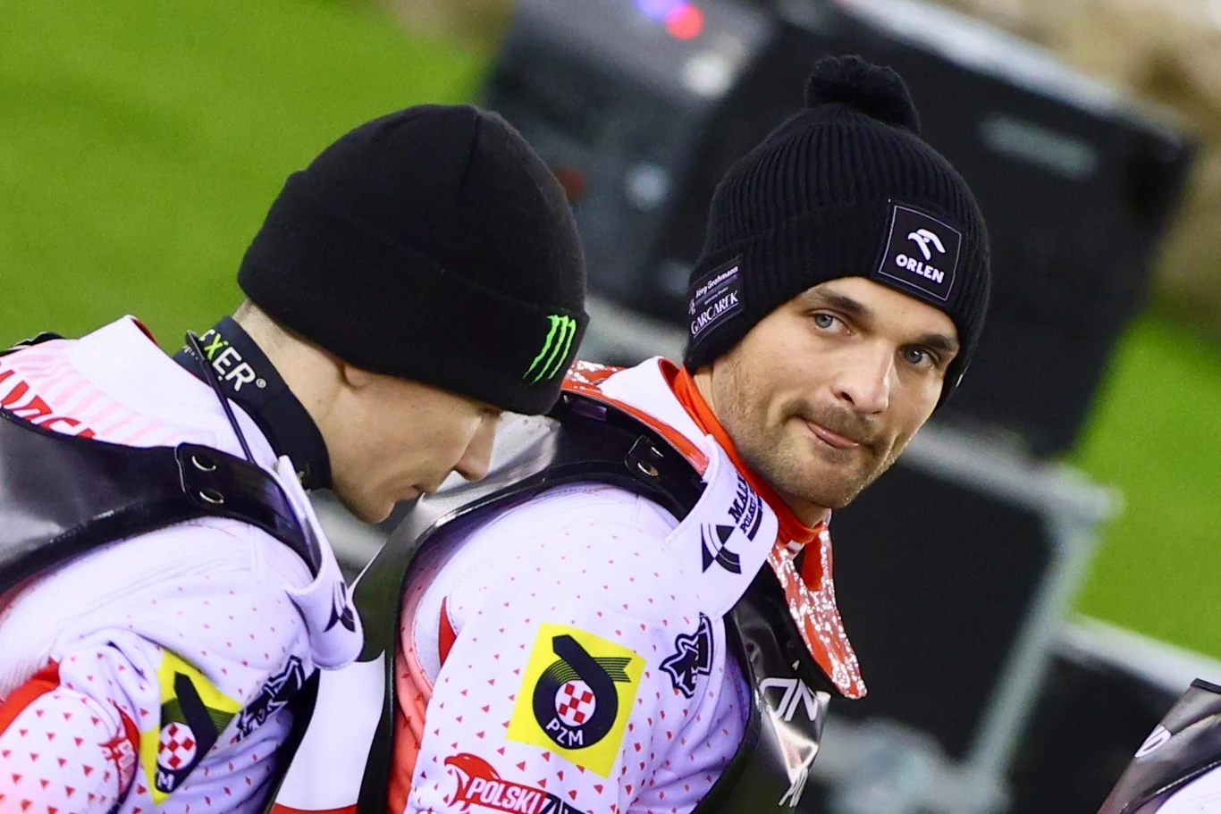 Dwóch żużlowców w białych strojach sportowych, ubrani w czarne czapki, stoją obok siebie na stadionie, na ich kombinezonach widoczne są logotypy sponsorów, jeden z nich patrzy w stronę obiektywu.