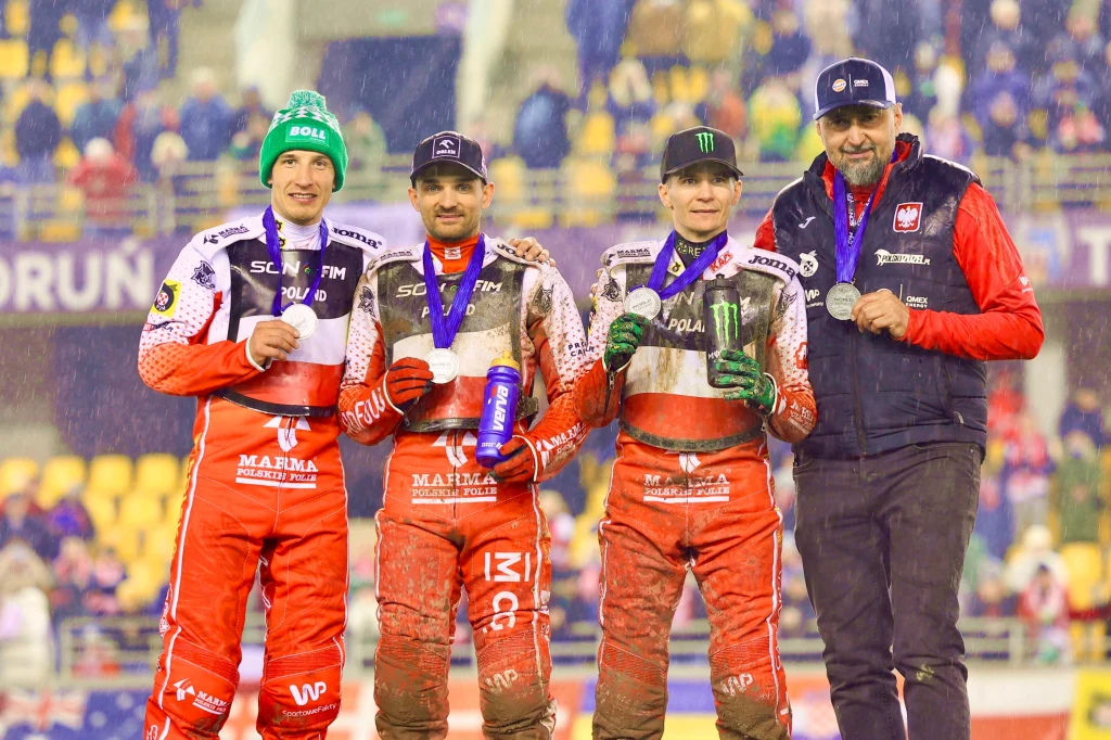 Od lewej: Piotr Pawlicki, Bartosz Zmarzlik, Patryk Dudek i Rafał Dobrucki Czterech mężczyzn ubranych w sportowe kombinezony żużlowe i kurtki pozuje razem na tle trybun stadionu, trzymając medale i uśmiechając się po zakończonych zawodach.