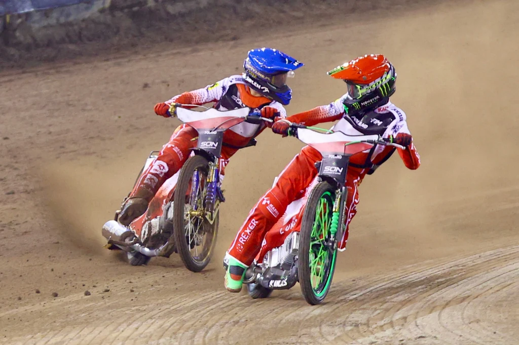 Patryk Dudek (czerwony kask) i Bartosz Zmarzlik (niebieski kask) Dwóch żużlowców w kombinezonach wyścigowych rywalizuje na torze, motocykle pochylone w trakcie dynamicznego zakrętu, wokół unosi się kurz, zawodnicy skoncentrowani na walce o pozycję.