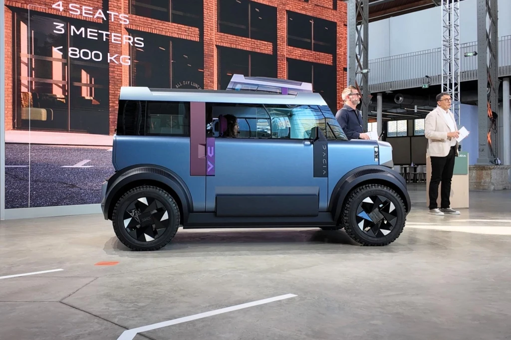 Dacia Hipster Concept mierzy równo 3 metry, co oznacza, że jest o 70 cm krótsza od Springa. Nowoczesny, kompaktowy samochód koncepcyjny o futurystycznym, pudełkowatym kształcie, stojący w przestrzeni wystawienniczej, obok dwóch osób prowadzących prezentację.