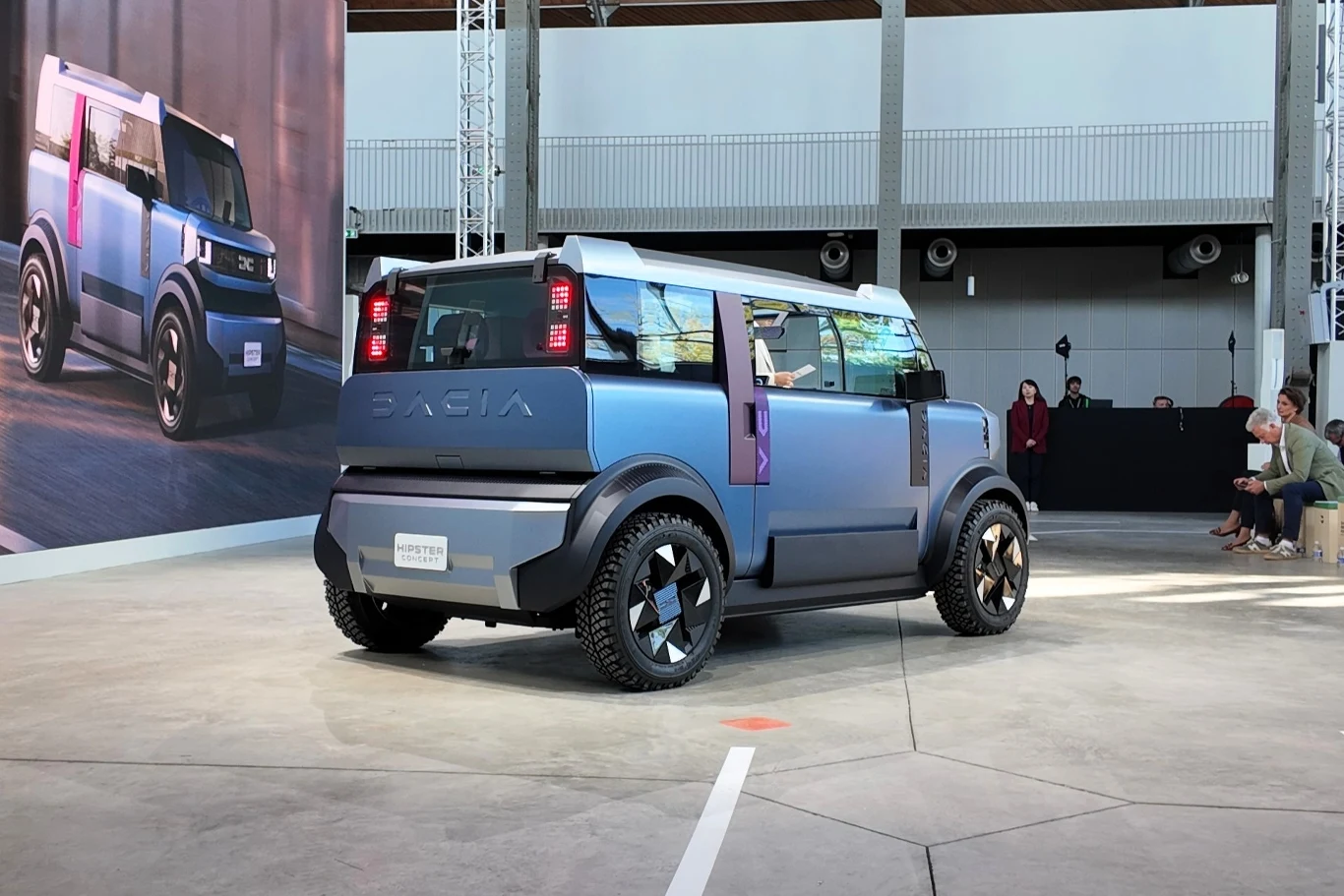Tył Dacii Hipster Concept został zaprojektowany z myślą o funkcjonalności. W efekcie klapa bagażnika jest dzielona, by ułatwić dostęp. Nowoczesny, elektryczny samochód koncepcyjny w kolorze niebieskim o futurystycznym designie, ustawiony w przestronnej hali wystawowej z betonową podłogą; w tle widoczny fragment dużego wydruku z tym samym pojazdem oraz kilka osób siedzących na ławkach.