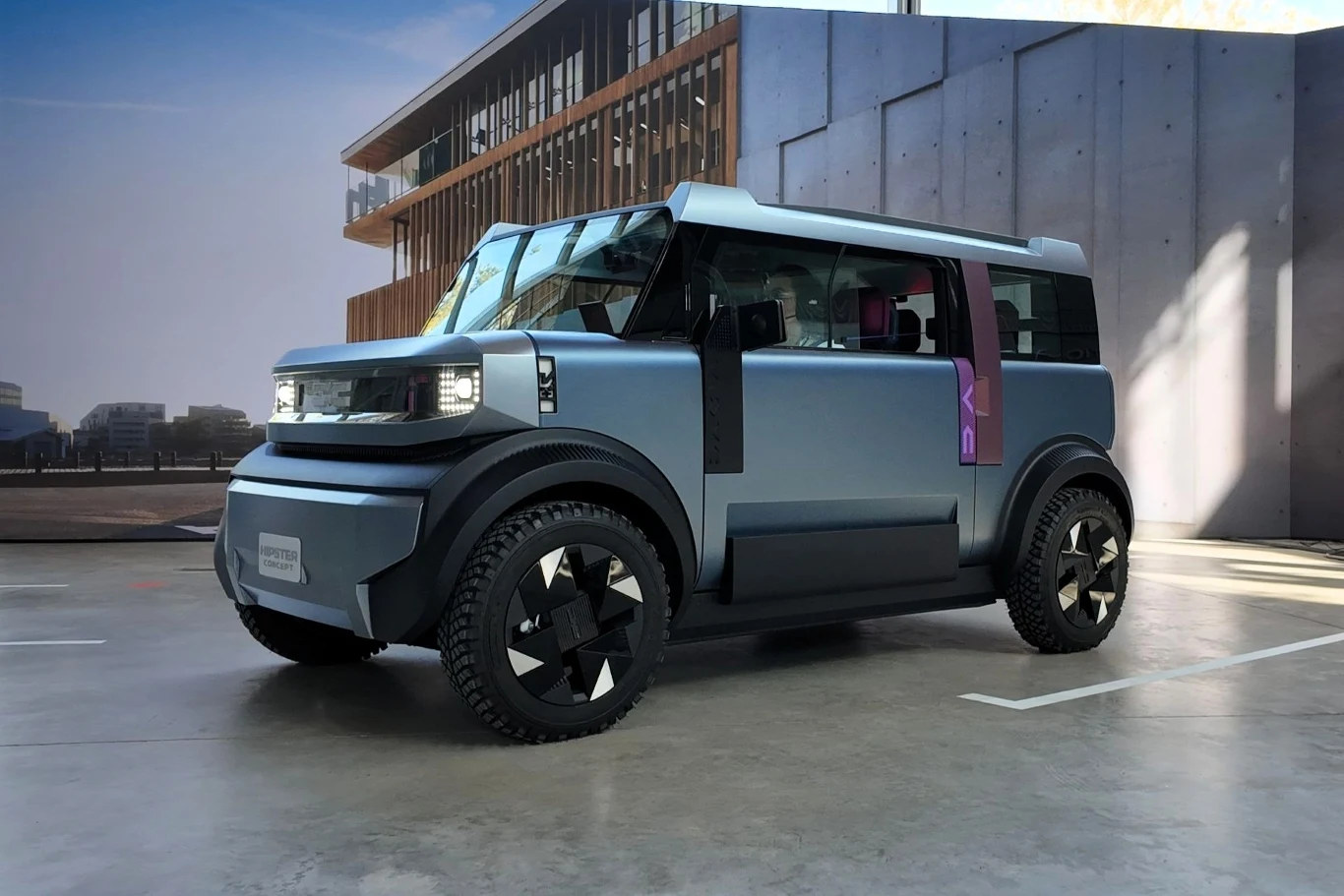 Dacia Hipster Concept to samochód elektryczny zapewniający zasięg do codziennych przejazdów z dwoma ładowaniami w tygodniu. Nowoczesny, kompaktowy samochód koncepcyjny o surowych, geometrycznych liniach nadwozia w matowym, niebieskoszarym kolorze, stoi na betonowej powierzchni w pobliżu przeszklonego budynku i betonowej ściany.