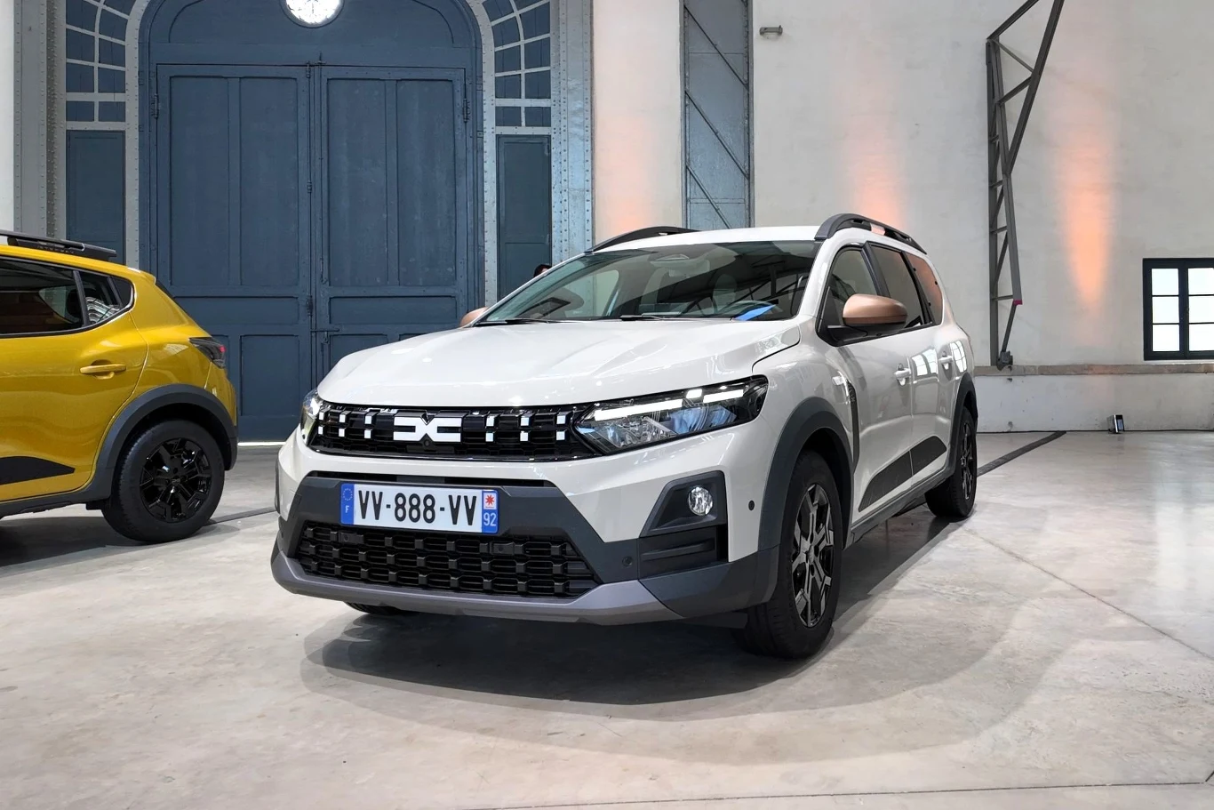 Aż 75 proc. sprzedanych w 2025 roku w Polsce nowych Dacii Jogger wyposażonych było w fabryczną instalację gazową Biały samochód osobowy marki Dacia Jogger zaparkowany w nowoczesnym, przestronnym wnętrzu, w tle widoczny fragment żółtego samochodu i duże, niebieskie drzwi.