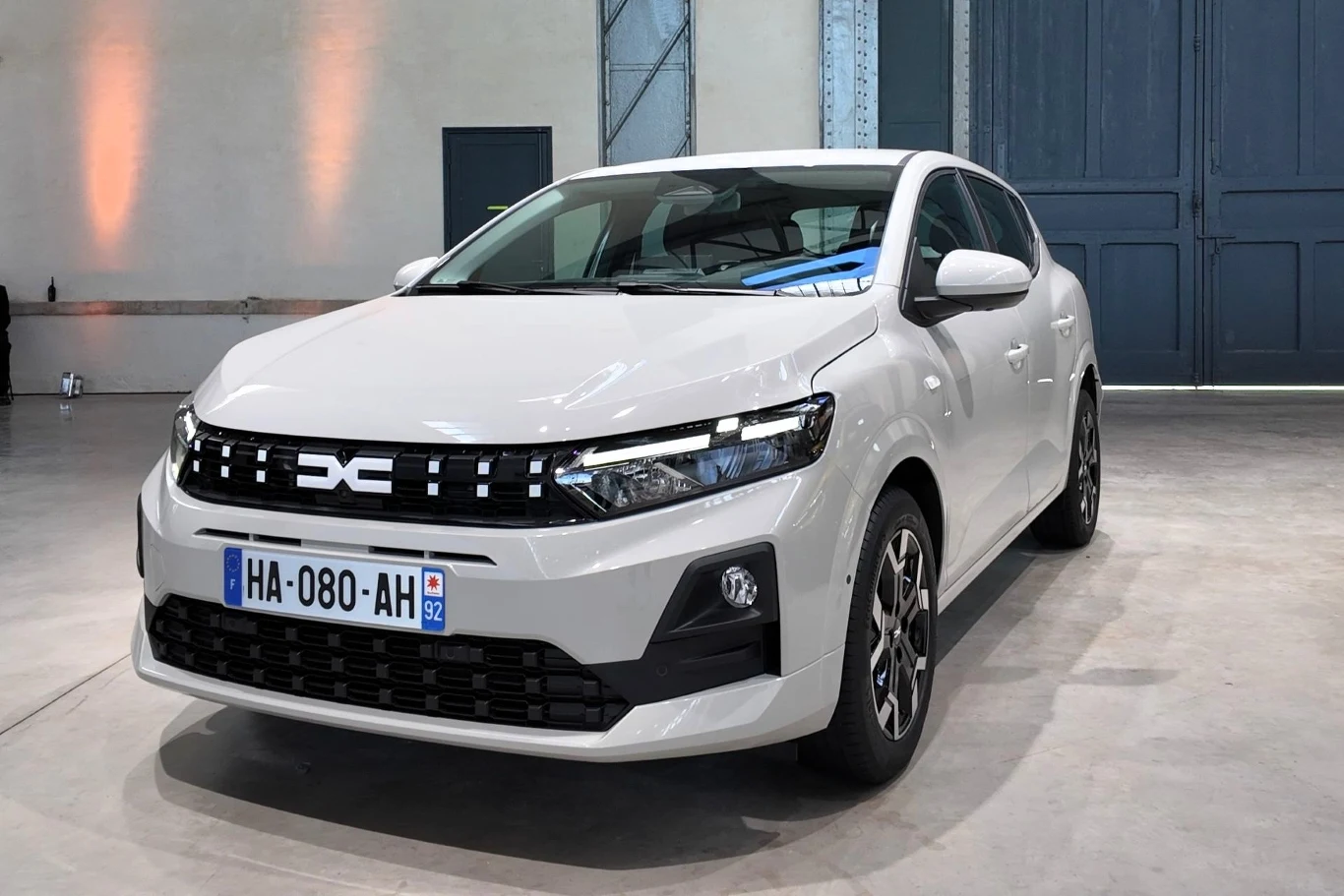 Biały samochód marki Dacia stoi w jasnym, industrialnym wnętrzu z betonową podłogą i dużymi drzwiami w tle; nowoczesny design przedniej części, widoczne światła LED oraz tablica rejestracyjna francuska.