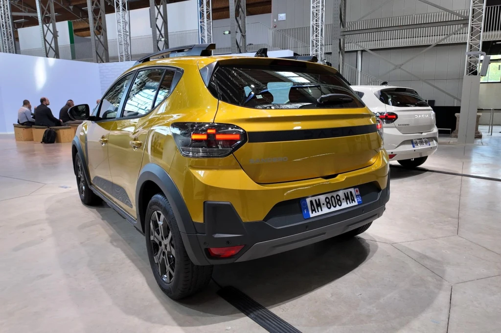 W Dacii Sandero Stepway, oprócz nowych świateł pojawił się również przeprojektowany tylny zderzak i nową listwę w kolorze matowej czerni biegnącą między światłami. Nowoczesny, żółty SUV zaparkowany w hali wystawowej, widok od tyłu, w tle biały samochód, metalowe konstrukcje i osoby siedzące na ławkach.