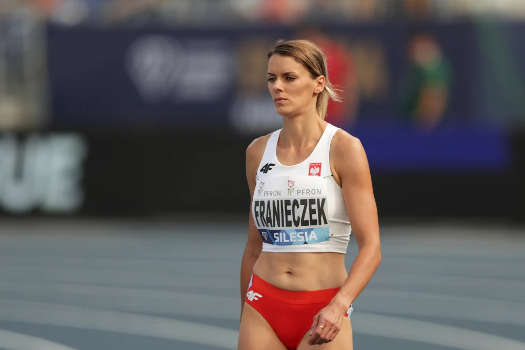 Justyna Franieczek Lekkoatletka w stroju sportowym z numerem startowym na piersiach, stojąca na bieżni stadionu sportowego, w tle rozmyte postacie i elementy trybun.