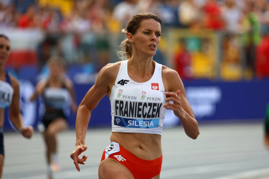 Justyna Franieczek Zawodniczka biegnąca na bieżni podczas zawodów lekkoatletycznych, ubrana w strój sportowy z numerem startowym i nazwiskiem, w tle inne sportsmenki oraz widownia