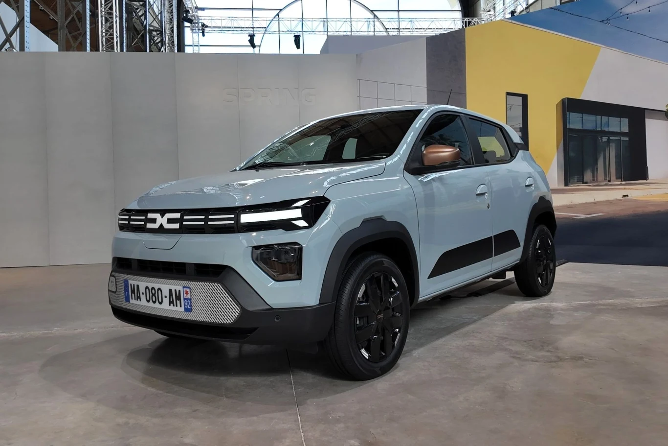 Jasnoniebieski samochód elektryczny marki Dacia Spring nowej generacji, zaparkowany w przestronnym, zadaszonym pomieszczeniu. Auto ma charakterystyczną czarną maskownicę, nowoczesne reflektory LED, czarne felgi i widoczne detale na zderzakach. W tle no...