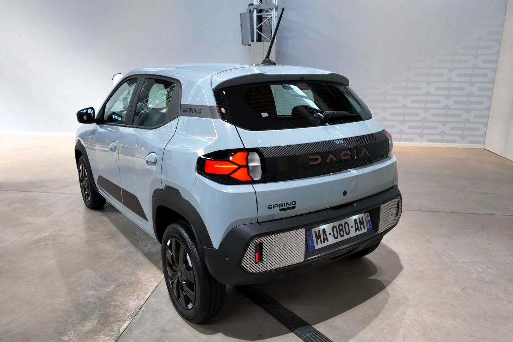 Dacia Spring dostępna będzie również z nowymi układami napędowymi. Zamiast wariantów o mocy 45 KM i 65 KM w ofercie znajdą się układy generujące 70 KM i 100 KM. Białosrebrny samochód Dacia Spring zaparkowany w jasnym pomieszczeniu na betonowej posadzce, widok z tyłu, dobrze widoczna tablica rejestracyjna oraz charakterystyczne tylne lampy pojazdu.